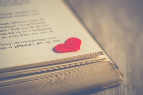 A photo of the corner of a page in an open book. A red heart, cut from paper, rests on the edge of the page.