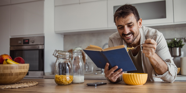 Man smiling while reading a book at his kitchen counter and eating cereal, hopefully not too close to the book because food stains in books are not cool. 