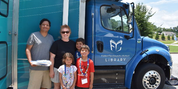 A family with three kids stands in front of the FCPL Bookmobile, "Loanie." All three kids in the family are wearing summer reading lanyards with numerous achievement buttons. A family with three kids stands in front of the FCPL Bookmobile, "Loanie." All three kids in the family are wearing summer reading lanyards with numerous achievement buttons.