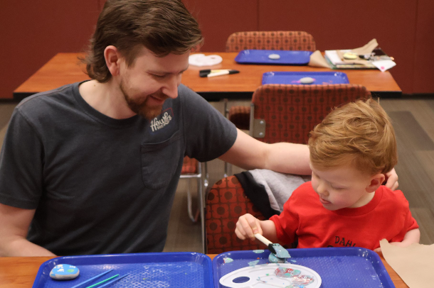 A proud, smiling father with his arm around his toddler son as the son intently paints a rock at Hampton Park Library. 