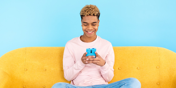 A smiling young man looking at his cell phone while sitting on a bright yellow sofa. 