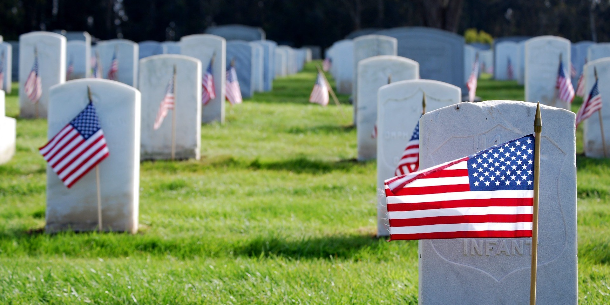 Rows of white marble headstones in a military cemetery, each adorned with a small American flag planted in the green grass.