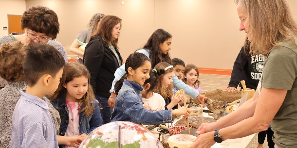 A group of smiling, interested children and adults exploring a table of objects in a program titled Lessons from the Cherokee with Elachee Nature Center. 