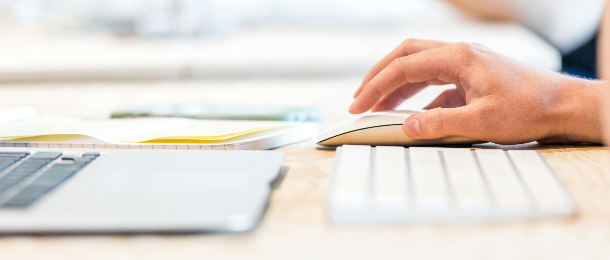 Close up of a hand using a computer mouse next to a keyboard and laptop computer. 