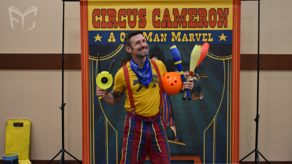 Circus Cameron holding props while standing in front of a sign that says Circus Cameron A One Man Marvel. 