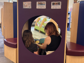An older child reads with a younger child in a reading nook.