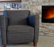 A photograph of a comfortable arm chair, next to a shelf of books and an electric fireplace.