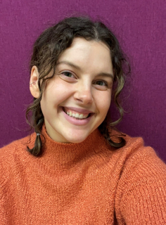A headshot picture of new Teen Librarian Holly in front of a purple background. She is smiling and has curly brown hair in 2 braids that come to her shoulders. She is wearing an orange sweater.