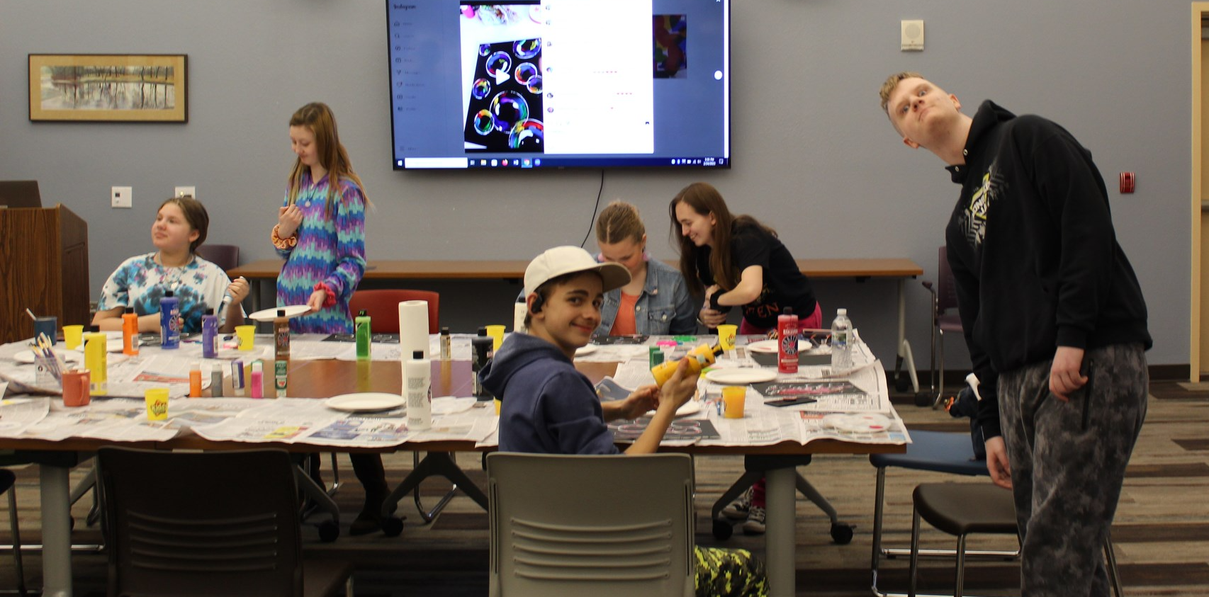 Image of six teenagers painting at a table. Several are making faces at the camera. 
