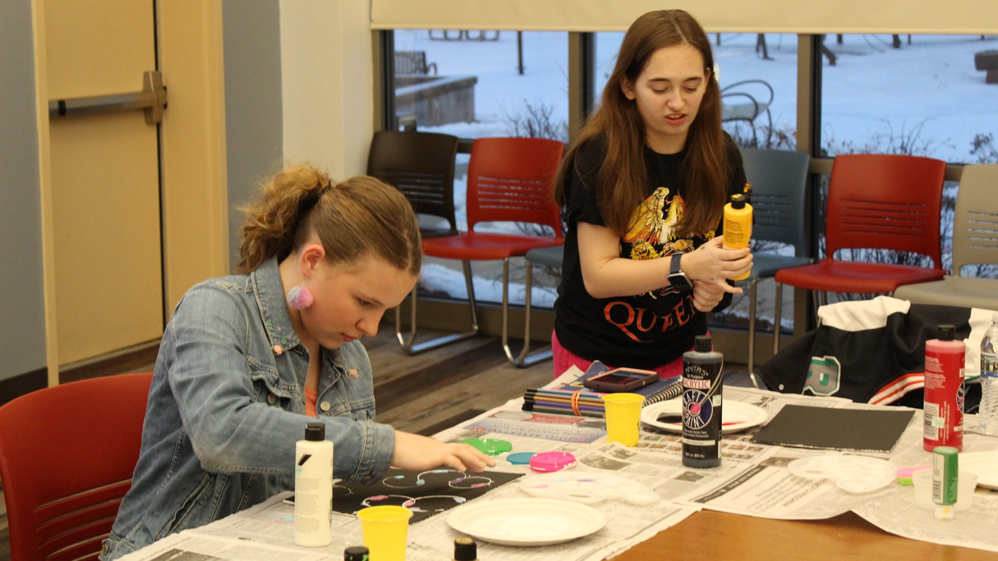 Image of two teen girls painting at a table. 