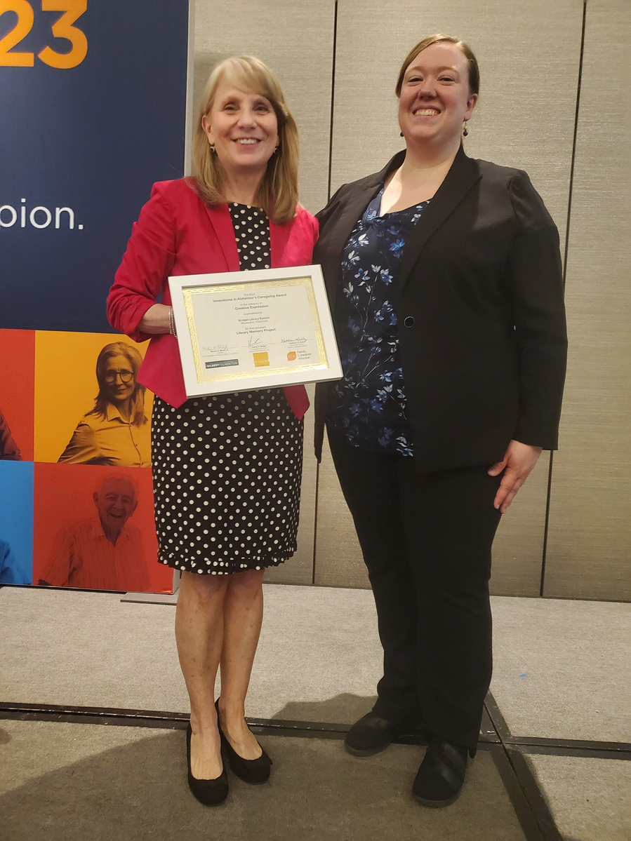 Karol Kennedy with Bridges Library System and Jennie Fidler with Oconomowoc Public Library stand on a stage at the On Aging 2023 Conference with Karol holding the Innovation Award