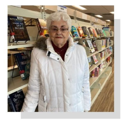 Georgi, an older woman with white hair and brown skin, wearing a white coat and glasses and standing in front of bookshelves at the Watertown Public Library