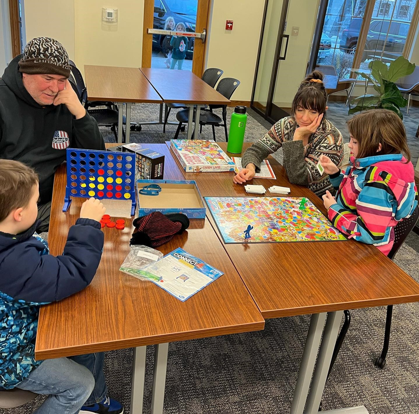 Two adults and two children sit at tables at the Hartland Public Library playing Connect 4 and another board game.