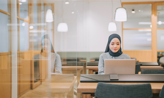 Photo of a woman in a hijab sitting at a laptop computer at a table in a library. Photo of a woman in a hijab sitting at a laptop computer at a table in a library.
