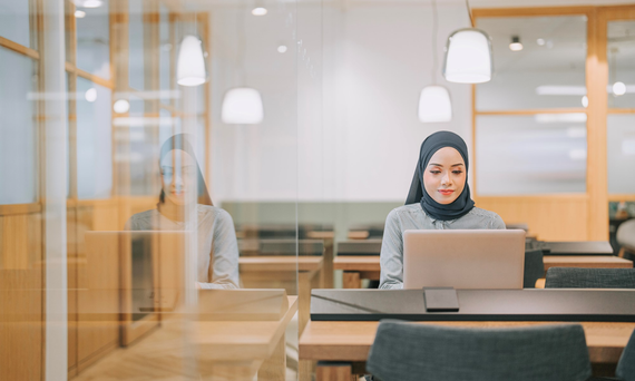 Photo of a woman in a hijab sitting at a laptop computer at a table in a library.