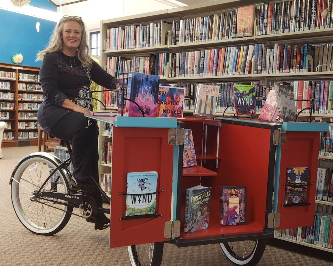Photo of Powers Memorial Library Director, Kristine Dexheimer, sitting on the book bike inside the library.