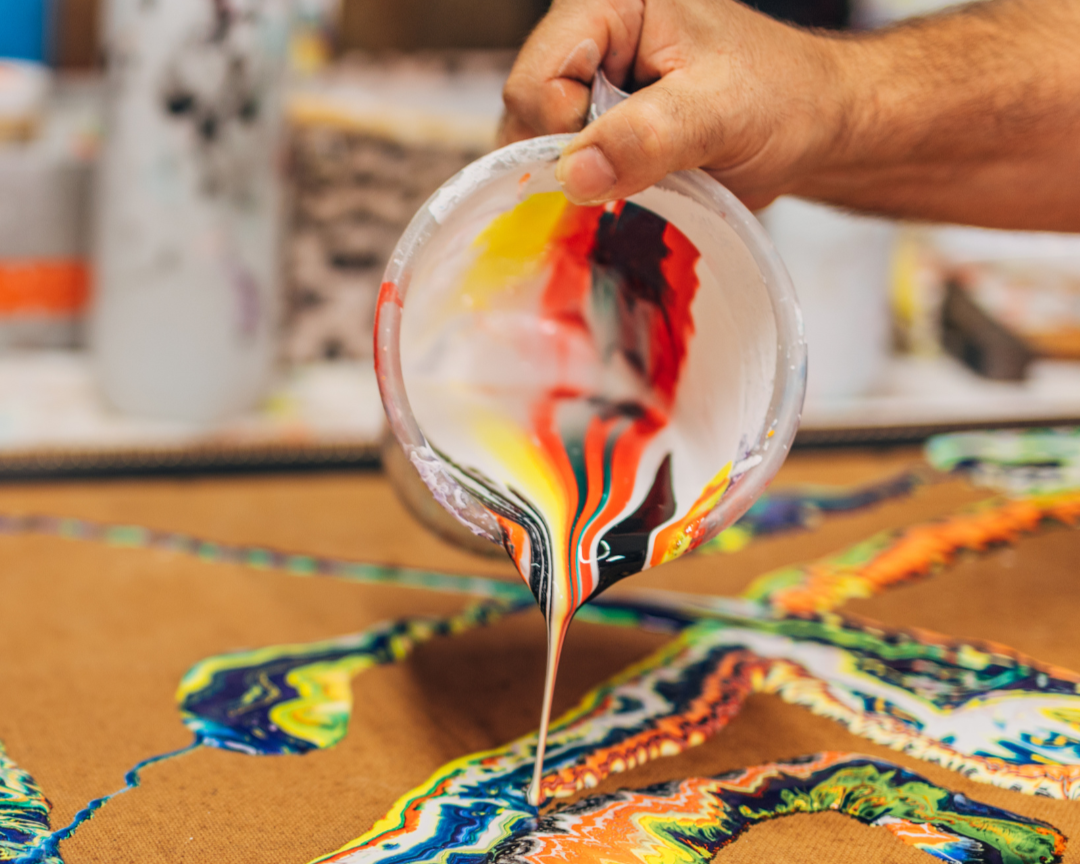 Photo of hand pouring mixed colors of paint from a small pitcher onto a board.