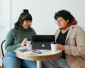 Stock photo of two people sitting together at a table indoors with coffee and a laptop computer.
