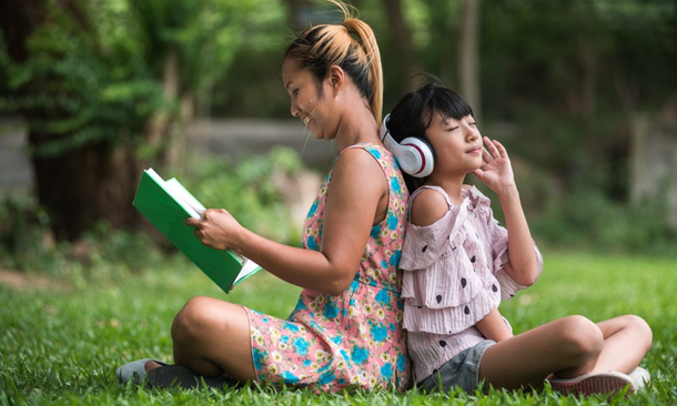 Woman Reading a Book and a Child with Headphones Sitting on Grass in a Park